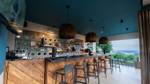 Un bar moderno con mostrador de madera, taburetes y luces colgantes. Hay estanterías con botellas y vasos, y vistas al océano a través de amplias ventanas.
