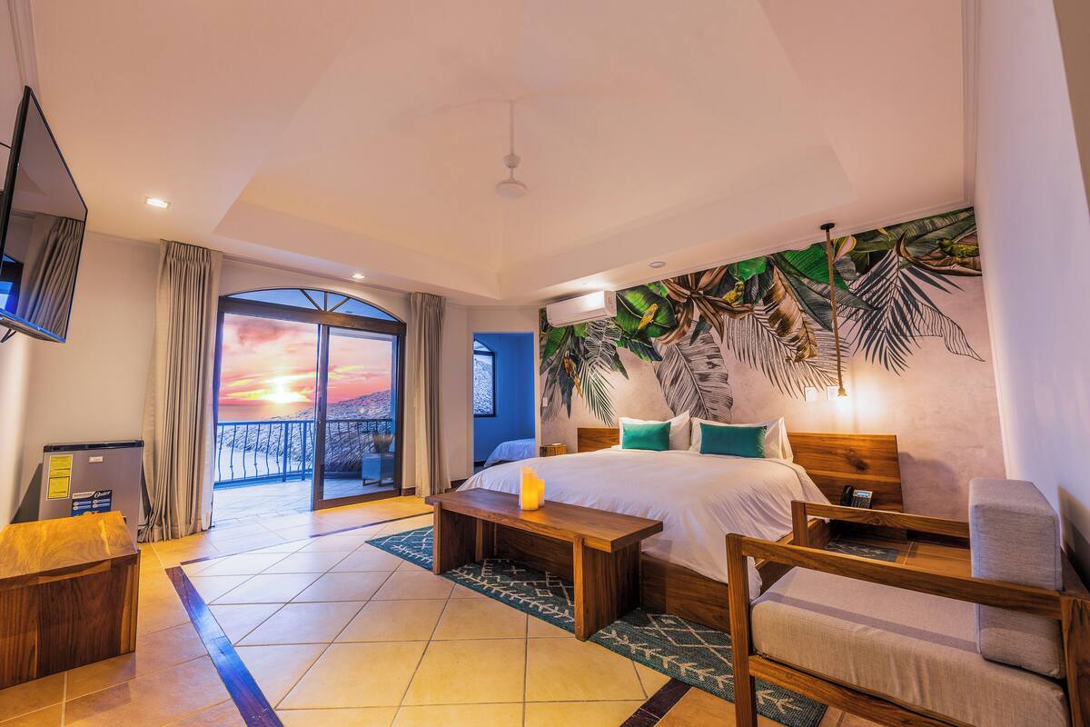 A hotel room with a bed, a wooden bench, a bedside table, a TV, and a view of the sunset through the balcony doors.
