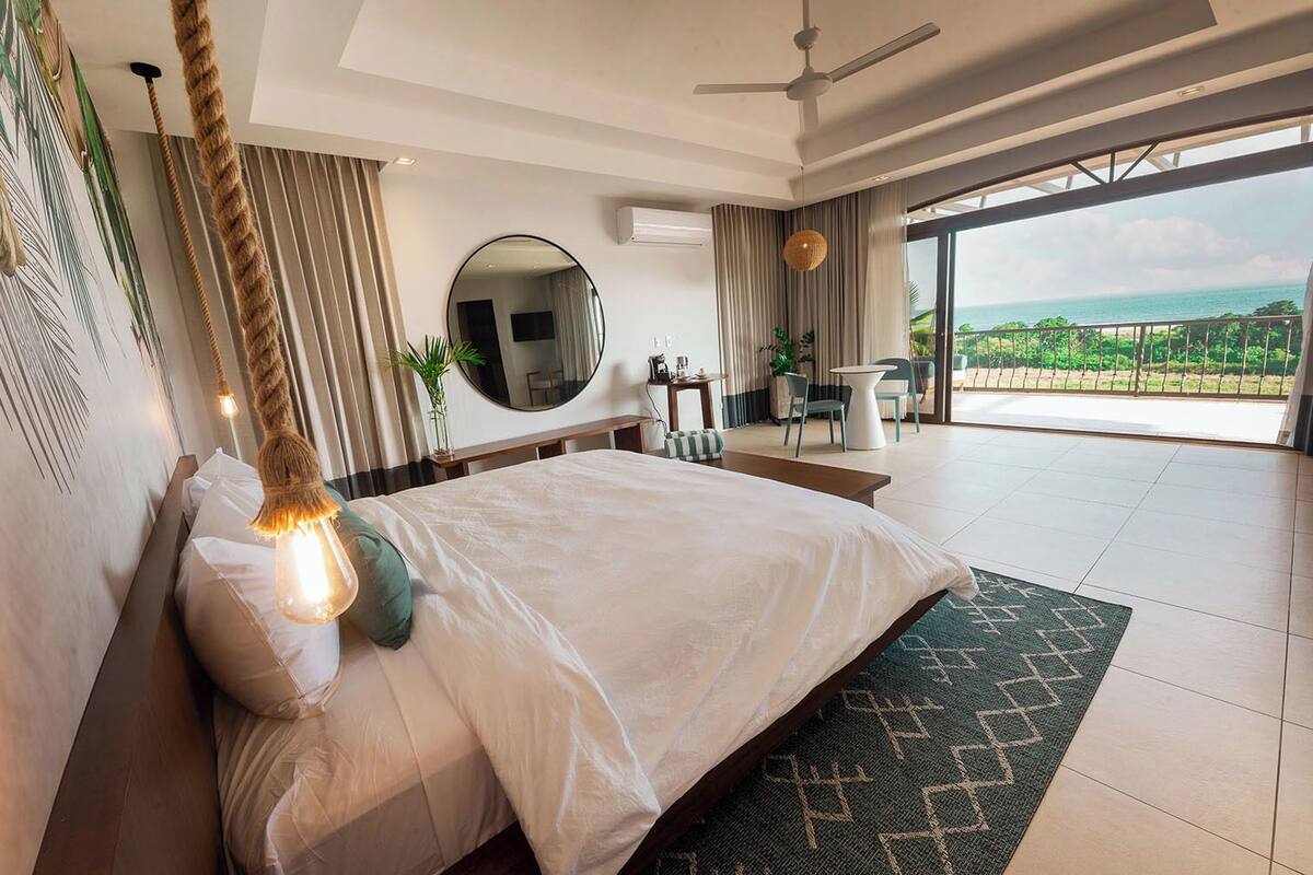 A modern hotel bedroom with a large bed, a ceiling fan, a view of the ocean, and a balcony with outdoor seating.
