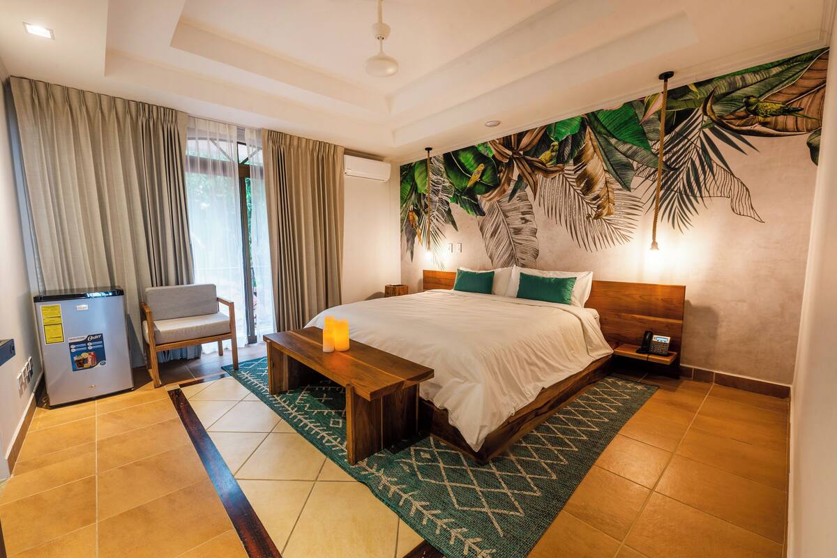 A hotel room with a bed, a bench, a chair, a nightstand, a wall-mounted phone, and a refrigerator.