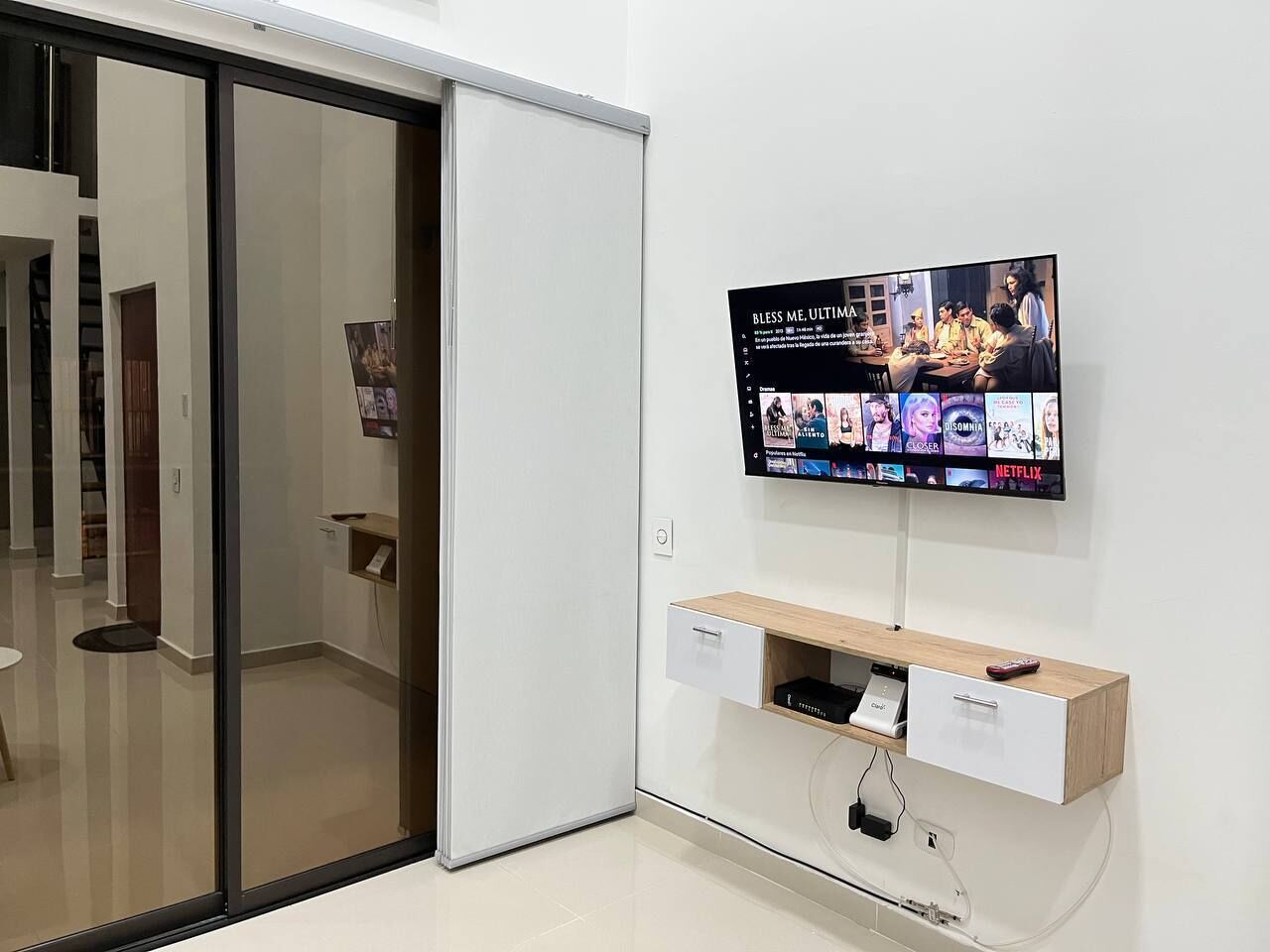 Una habitación moderna con una puerta corredera, un televisor montado que muestra varios programas, un estante de madera con un control remoto y un mueble cajonero blanco.