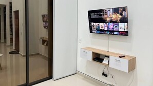 Una camera moderna con una porta scorrevole, una TV a parete che mostra diversi programmi, una mensola in legno con un telecomando e un mobile contenitore bianco.
