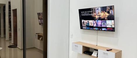 Una habitación moderna con una puerta corredera, un televisor montado que muestra varios programas, un estante de madera con un control remoto y un mueble cajonero blanco.