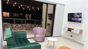 Un soggiorno moderno con un divano verde, una poltrona viola, un tavolino rotondo e un poggiapiedi in legno. Un televisore a schermo piatto è montato a parete, e si intravede un'area pavimentata in pietra attraverso il vetro della porta.