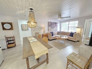 A spacious living room with a dining area, a sofa set, a chandelier, and a large window providing natural light.