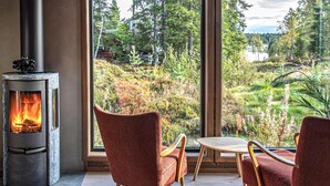 A cozy room with a wood-burning stove, two red chairs, a small table, and a large window offering a view of a forest and lake. - Beautiful home in Steinsholt with sauna (Steinsholt)