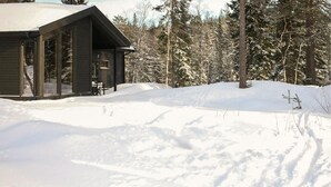 A cabin with a large window and a door, surrounded by snow-covered ground and trees. - Beautiful home in Steinsholt with sauna (Steinsholt)
