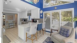 A modern kitchen with white cabinetry, a central island, and a dining area with bar stools. There is a large window with blinds and a view of greenery outside. - Beautiful Spacious Home in Palmetto Dunes w Pool 5 minute Walk to the Beach ! (Hilton Head Island)