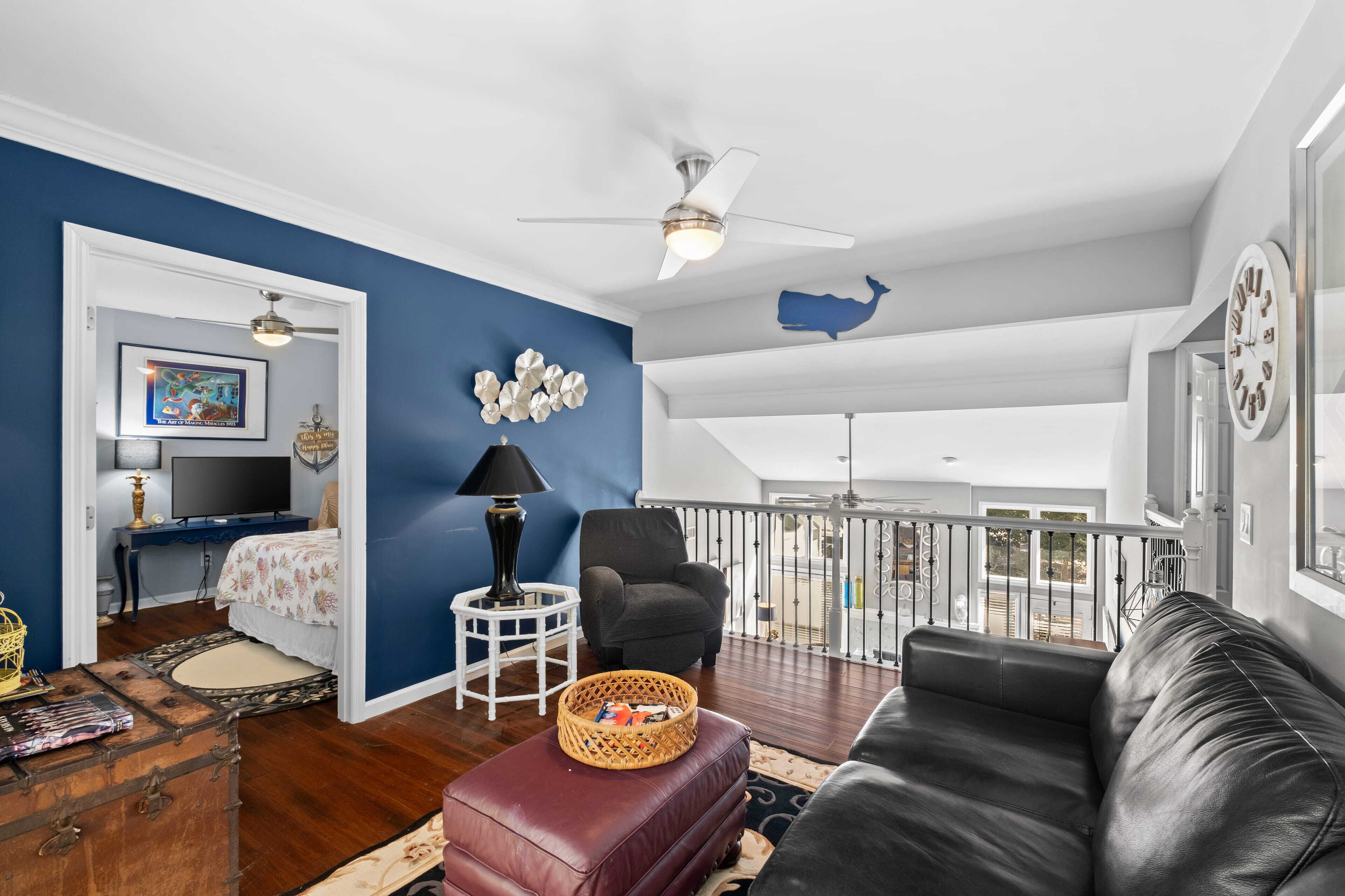 A spacious living room with a black leather couch, a red ottoman, and a blue accent wall. A ceiling fan and a large window provide natural light.