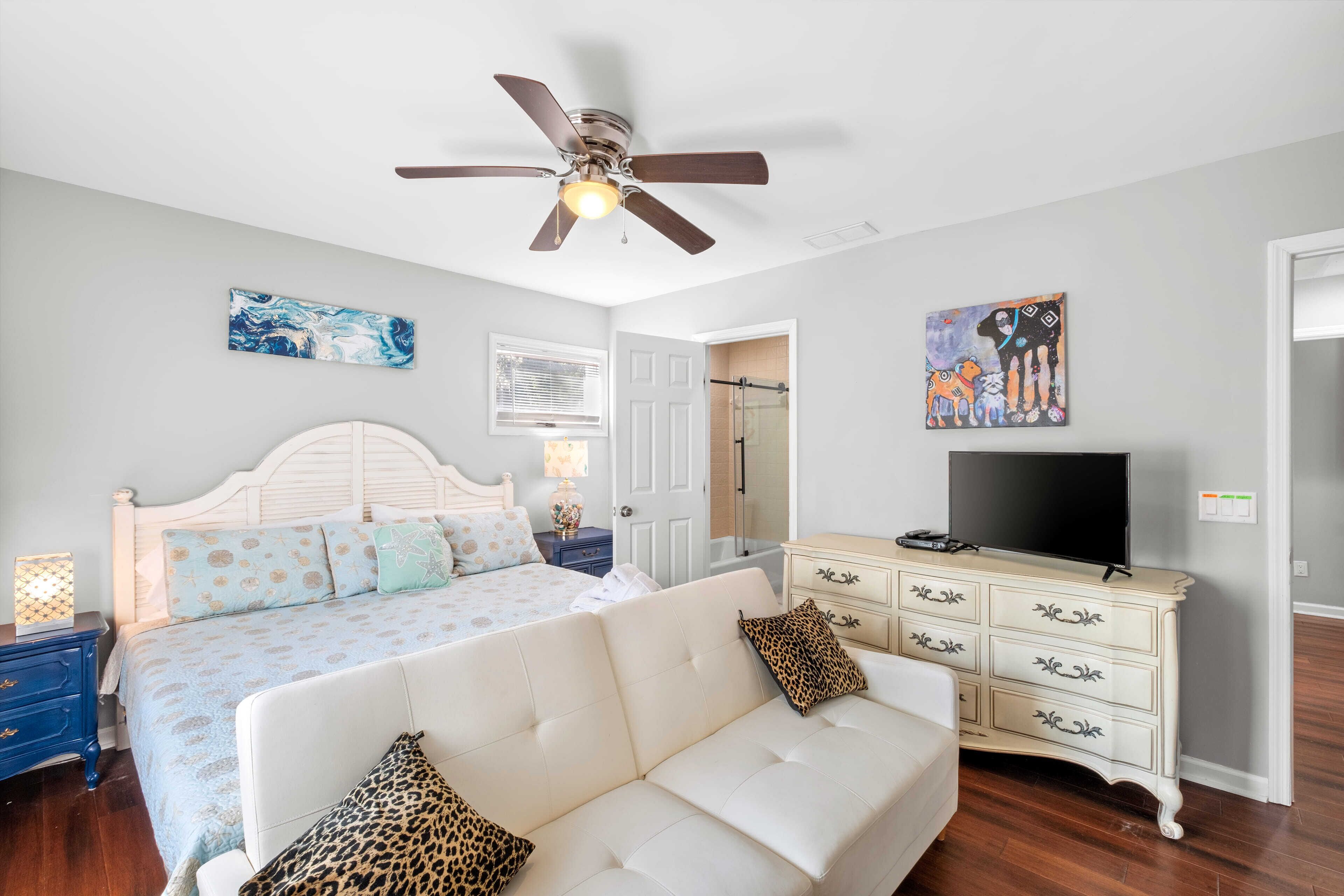 A bedroom with a bed, a white sofa, a TV, a dresser, and artwork on the walls.