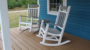 Deux fauteuils à bascule sur une véranda en bois, avec une maison bleue en arrière-plan.