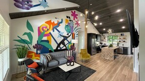 Un salon moderne avec une fresque murale abstraite aux couleurs vives, un canapé gris, un fauteuil jaune et une cuisine en arrière-plan.