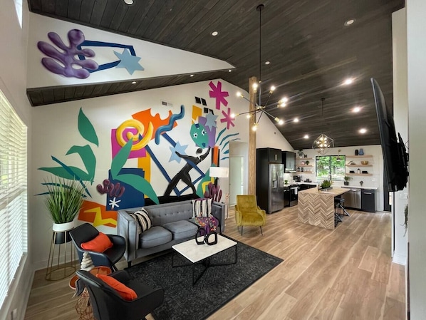 A modern living room with a colorful abstract mural, a gray sofa, a yellow armchair, and a kitchen in the background.