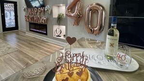 Une pièce moderne avec un plancher en bois, une cheminée, une table en verre sur laquelle se trouve un gâteau d’anniversaire, et une bouteille de vin.