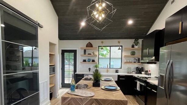 Een moderne keuken met een visgraat houten vloer, een centraal eiland, zwarte kastjes, een roestvrijstalen koelkast en een geometrische hanglamp.