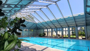 An indoor swimming pool with clear glass ceilings and large windows, surrounded by potted plants and lounge chairs.