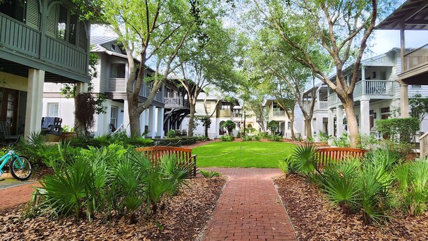 A courtyard with a brick pathway, surrounded by lush greenery and trees, leading to a well-maintained lawn.