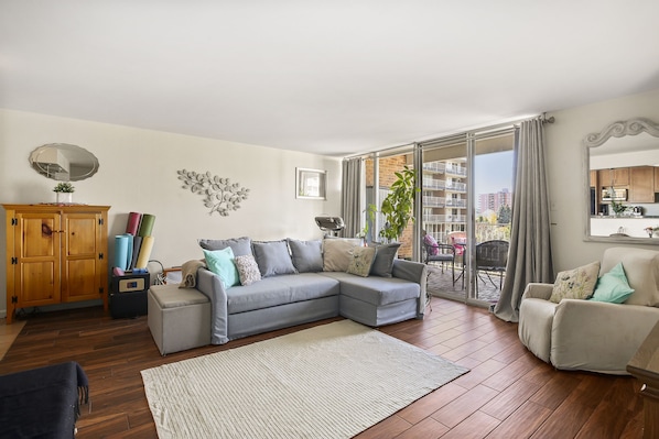 A living room with a grey sectional sofa, a wooden cabinet, a small table, and a large window with a view of buildings.