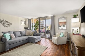 A living room with a grey sectional sofa, a white armchair, and a wooden cabinet. There is a large window with a view of a balcony and buildings. - 1 BED-Sunny Apt-Denver- Pool, gym, sauna, Walk to dining, shops, hospitals; (Denver)