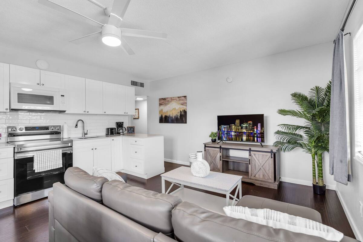 A modern kitchen with white cabinets, a microwave, and an oven. There is a dining area with a sofa and a table. A television is mounted on the wall.