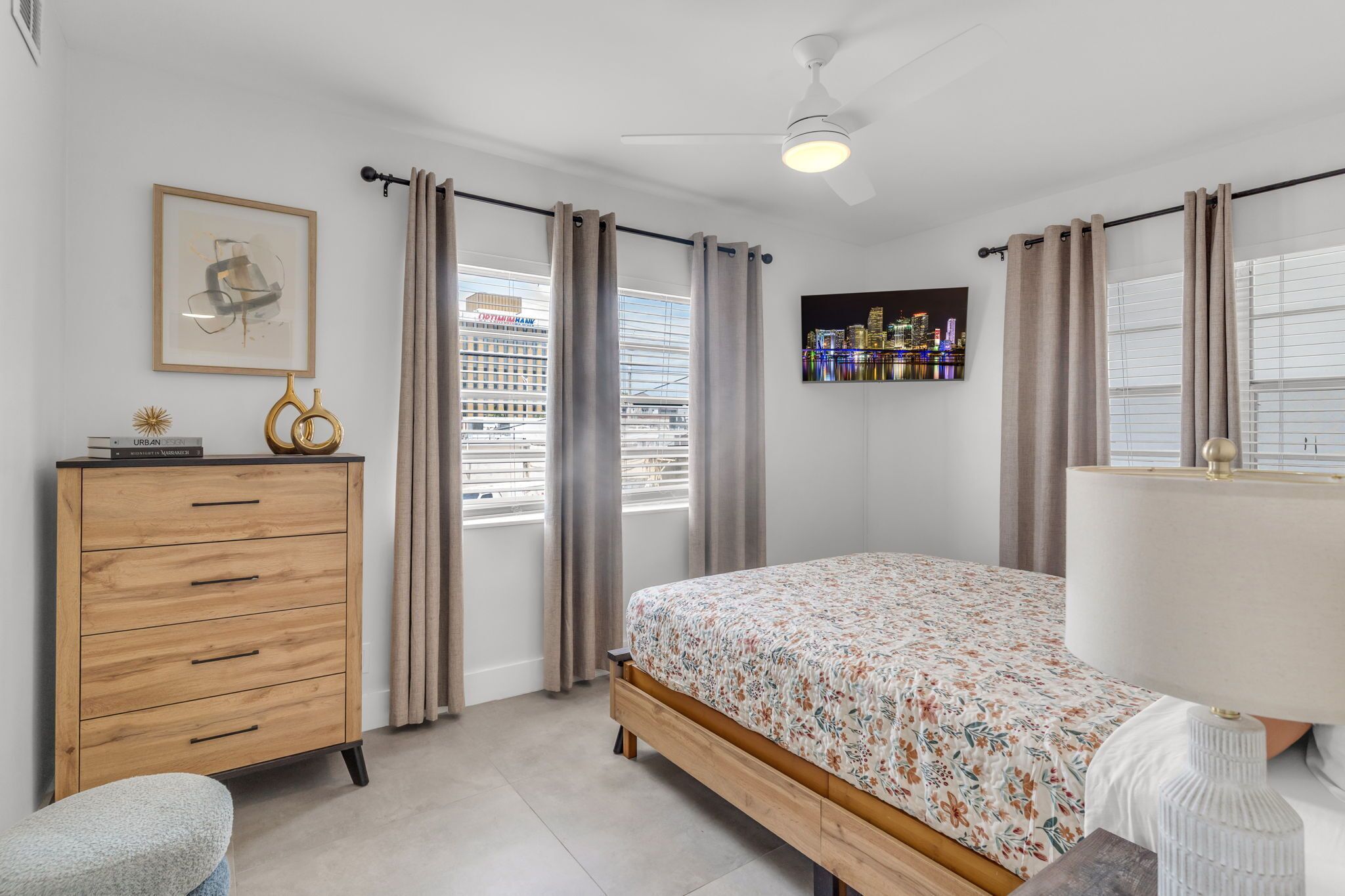 A bedroom with a bed, a dresser, a lamp, a ceiling fan, and a cityscape view through the window.