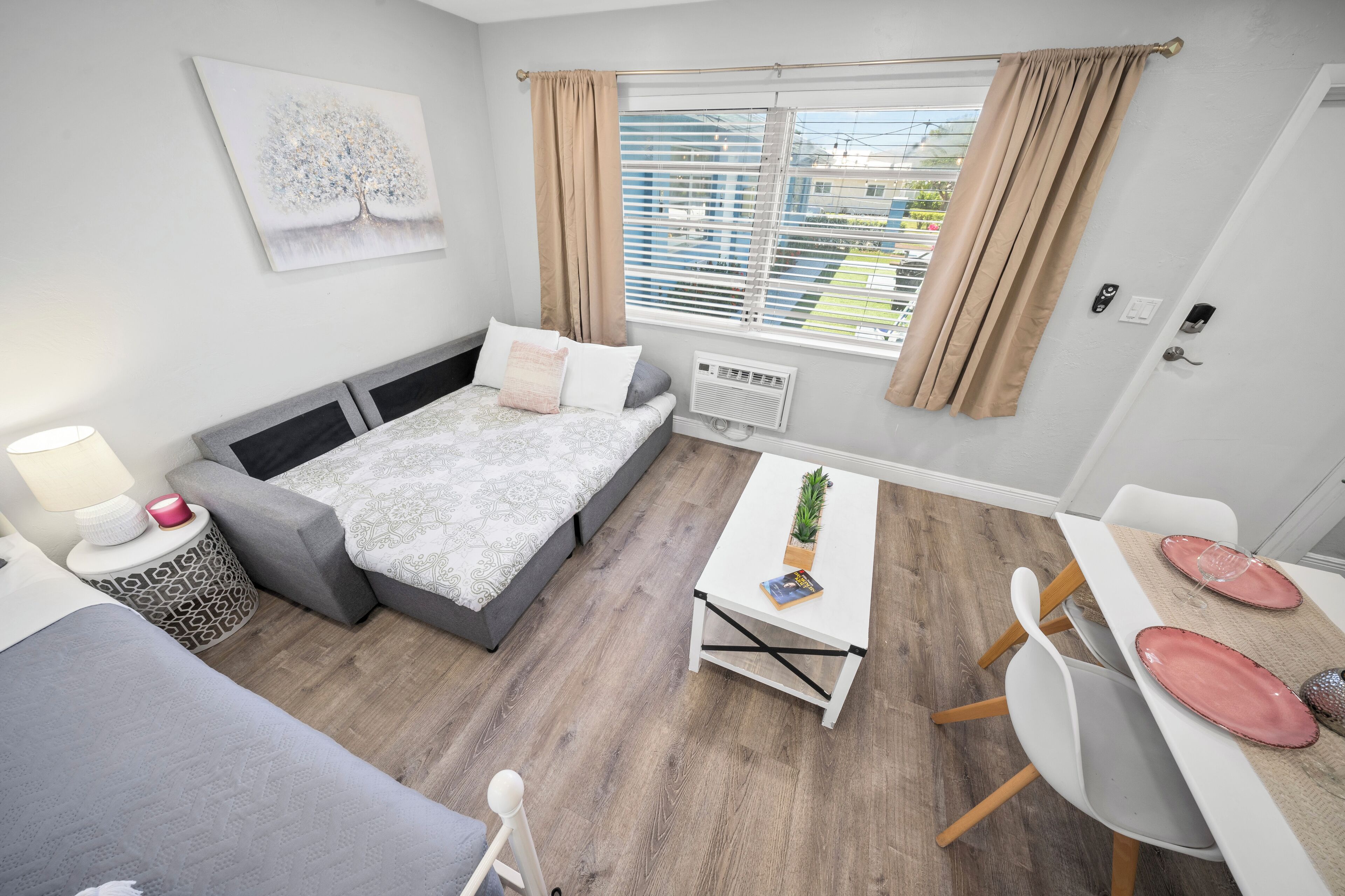 A modern, well-lit room with a bed, a small table with a plant, and a dining area with chairs and a table.