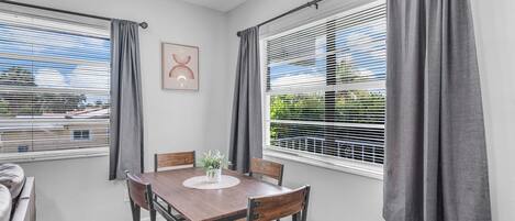 A dining area with a wooden table and chairs, a potted plant, and a view through window blinds.
