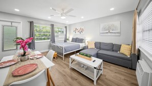 A modern living room with a sofa, a dining table set with plates and glasses, a ceiling fan, and a window with curtains. - Bayview Regency Apartments By Lowkl (Fort Lauderdale)