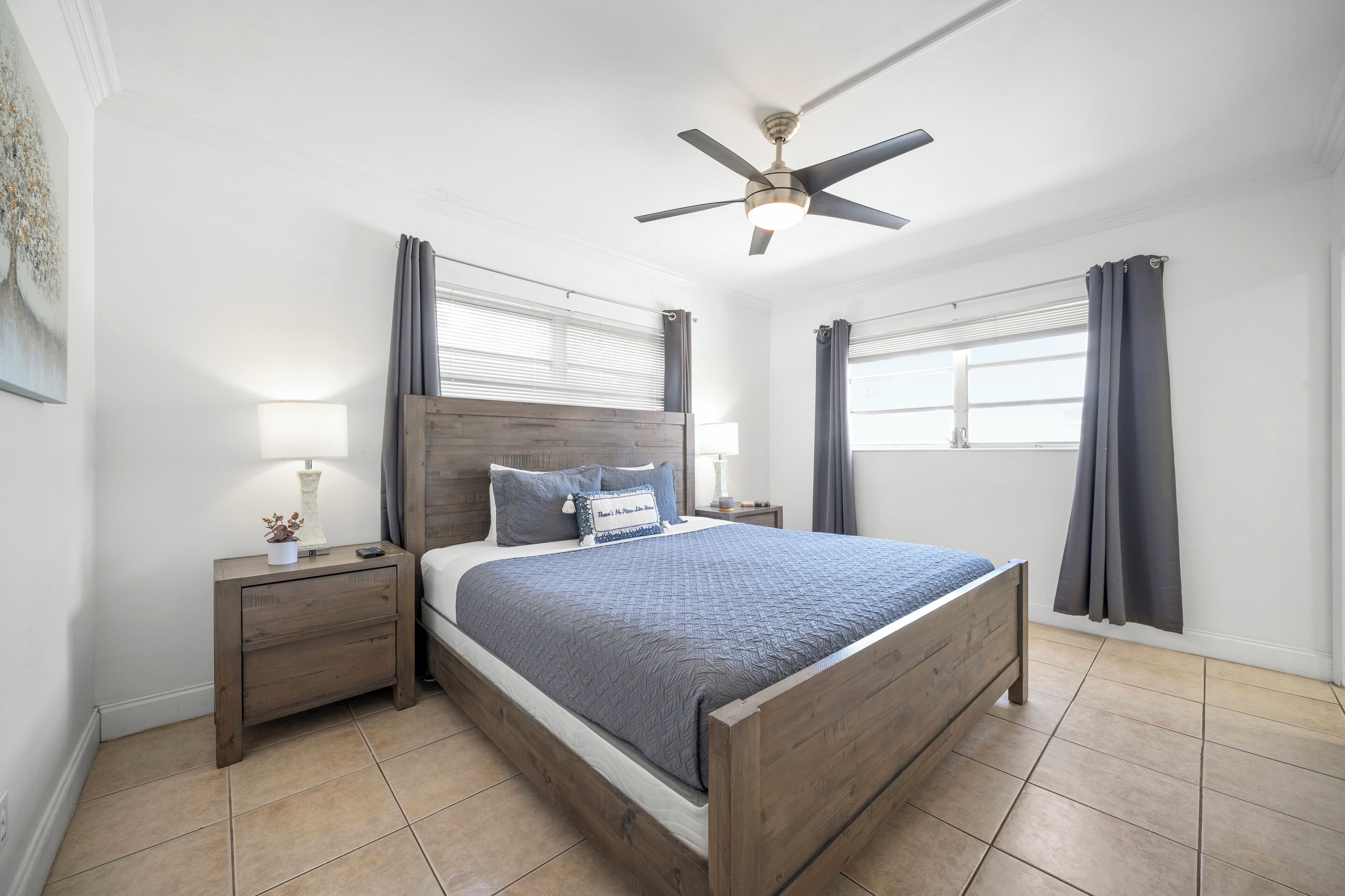 A bedroom with a wooden bed, two bedside tables with lamps, a ceiling fan, and windows with curtains.