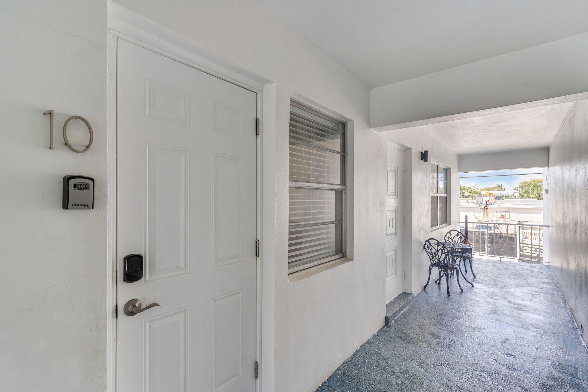 Un couloir avec une porte blanche, une fenêtre avec des stores, un petit espace extérieur aménagé avec des chaises et un balcon avec une balustrade.