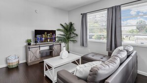A living room with a sofa, a coffee table, a TV, a plant, and a window with curtains. - Bayview Regency Apartments By Lowkl (Fort Lauderdale)