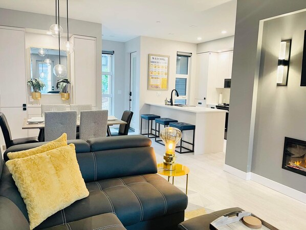 Una sala de estar moderna con un sofá de cuero negro, un cojín amarillo y una lámpara de mesa de vidrio.