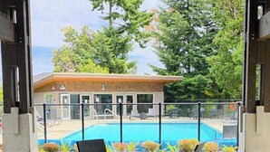 En uteplats med ett glasbord och flätade stolar, med utsikt över en swimmingpool och en byggnad med ett tak täckt av tegelpannor.