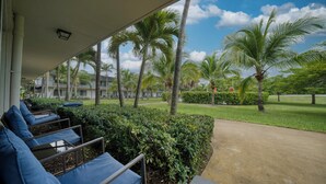 A covered outdoor seating area with blue cushioned chairs, palm trees, and a pathway leading to a building. - Riverwatch Apartments by Lowkl (Jensen Beach)