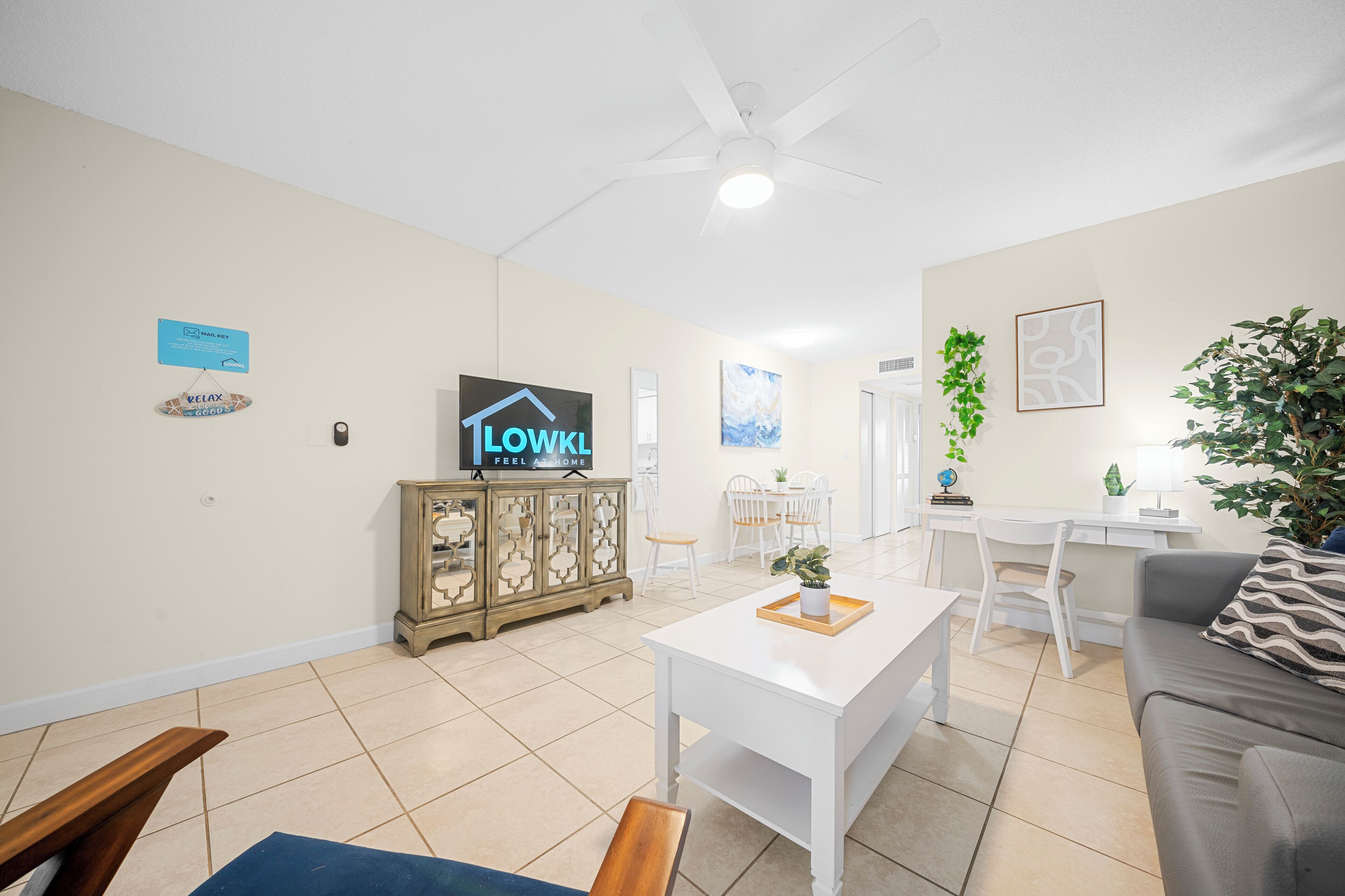 A spacious living room with a white sofa, a coffee table, and a TV on a cabinet. There is a dining area with a table and chairs, and a ceiling fan.