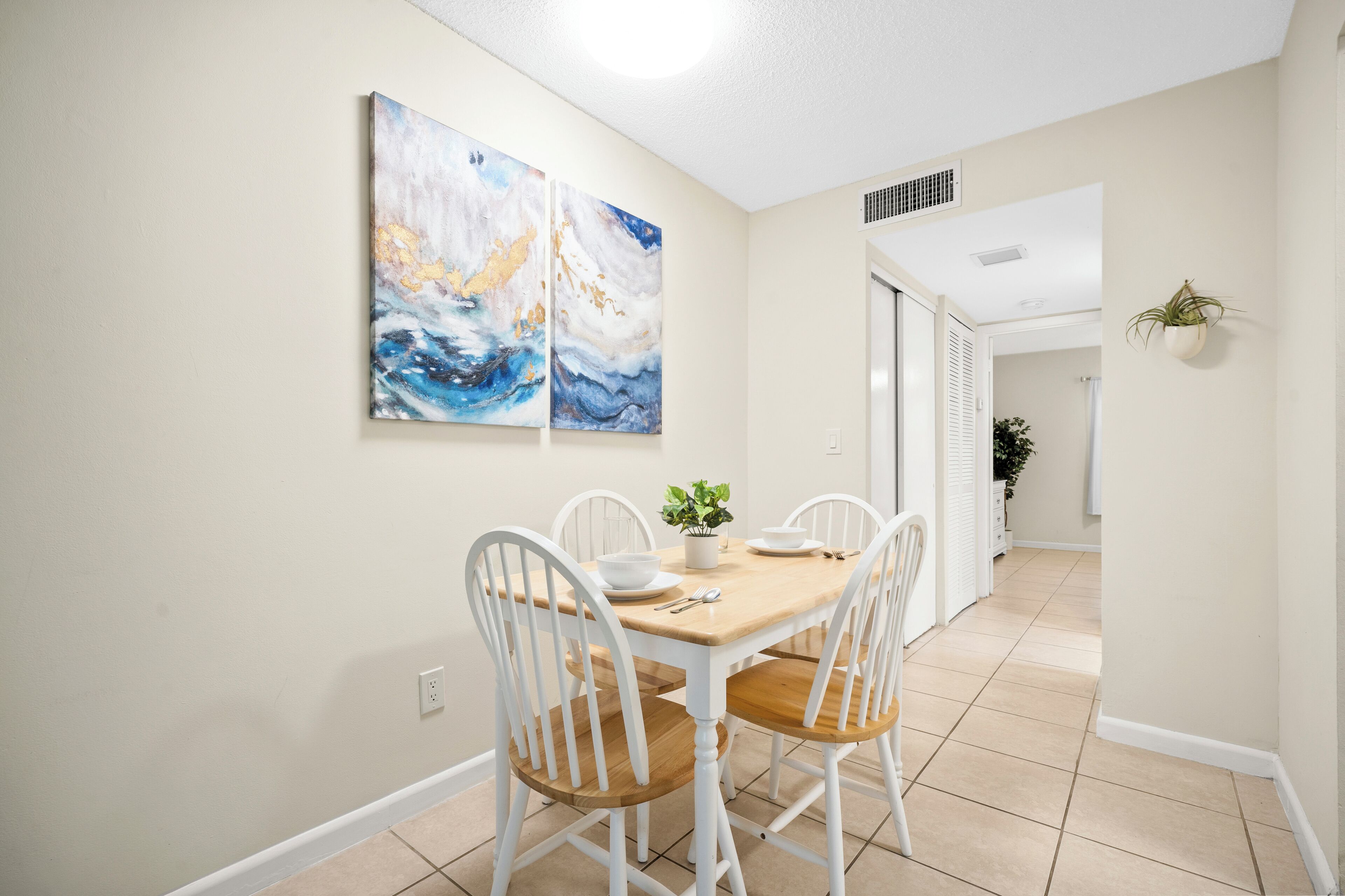 A dining area with a wooden table and white chairs, a potted plant, and two abstract paintings on the wall.