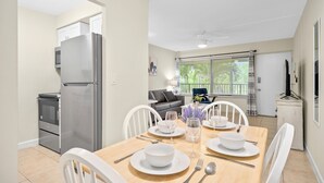 Comedor con mesa de madera dispuesta para cuatro personas, sillas blancas y zona de cocina con refrigerador y horno.