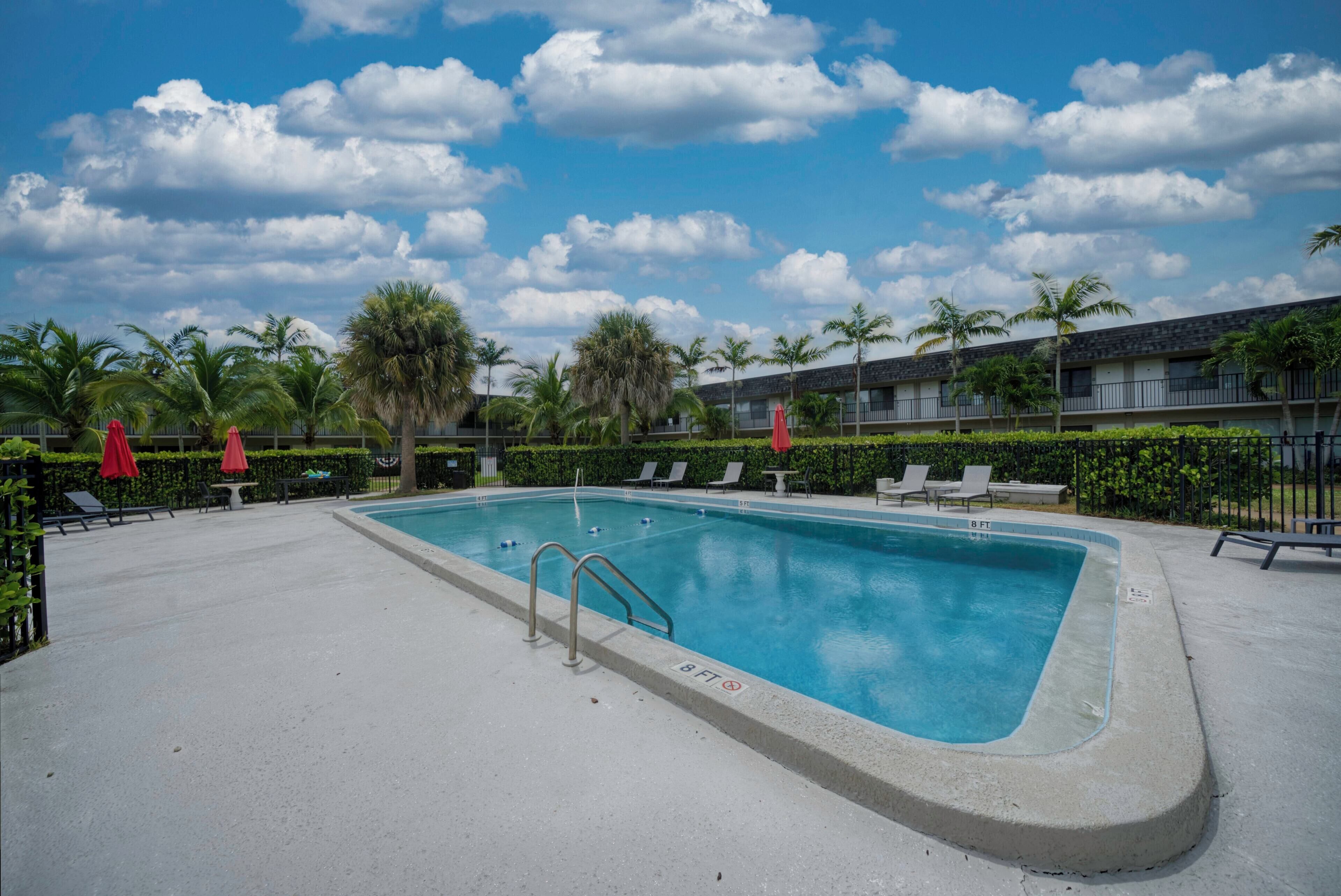 A rectangular swimming pool with a metal ladder, surrounded by lounge chairs and red umbrellas, with palm trees and a building in the background.