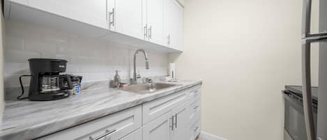 Una cocina con armarios blancos, encimera de mármol, cafetera y refrigerador de acero inoxidable.