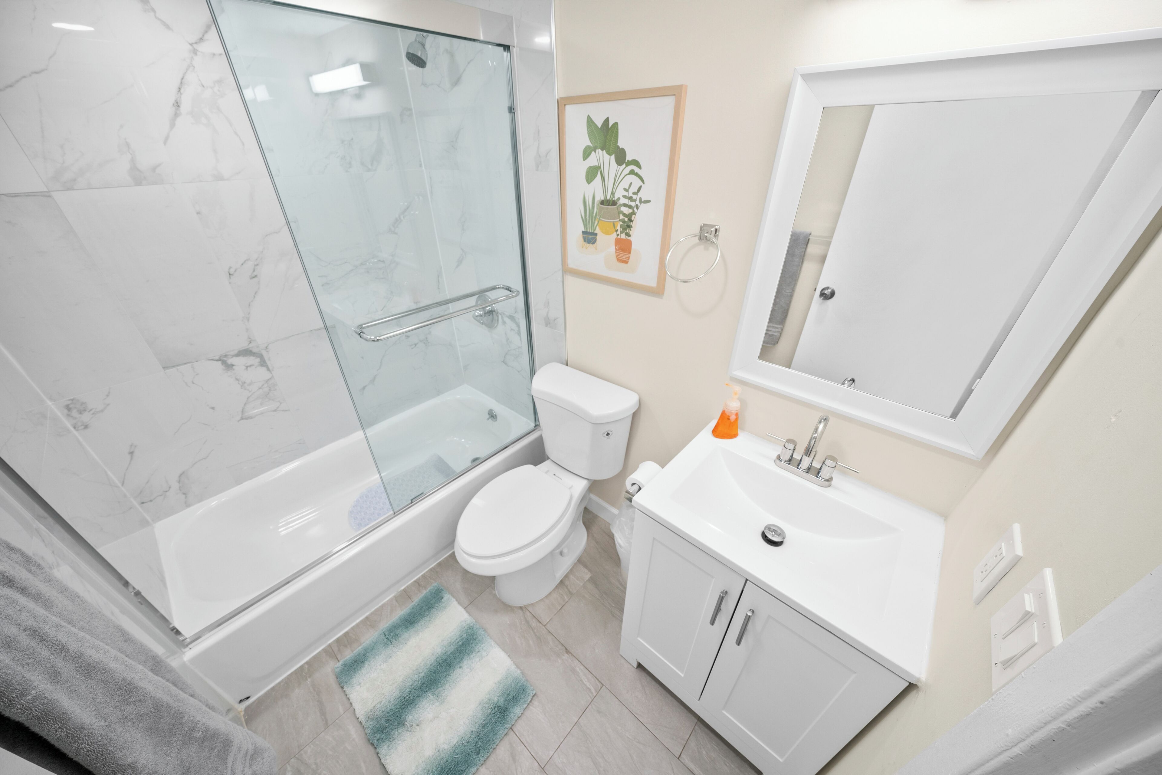 Une salle de bain moderne avec une douche à parois de verre, des toilettes, un lavabo avec un miroir et une plante décorative encadrée accrochée au mur.