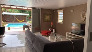 A modern living room with a grey sofa, a glass coffee table, a red ottoman, a TV on a white stand, and a sliding glass door leading to a pool area with a hammock and outdoor furniture. - Welcome to casa fall, a large family villa in the rainforest. Enjoyyyyy!!! (Fort-de-France)