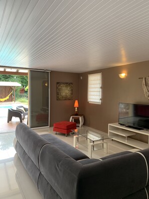 A modern living room with a grey sofa, a red ottoman, a glass coffee table, a TV on a white stand, and a wall-mounted lamp.