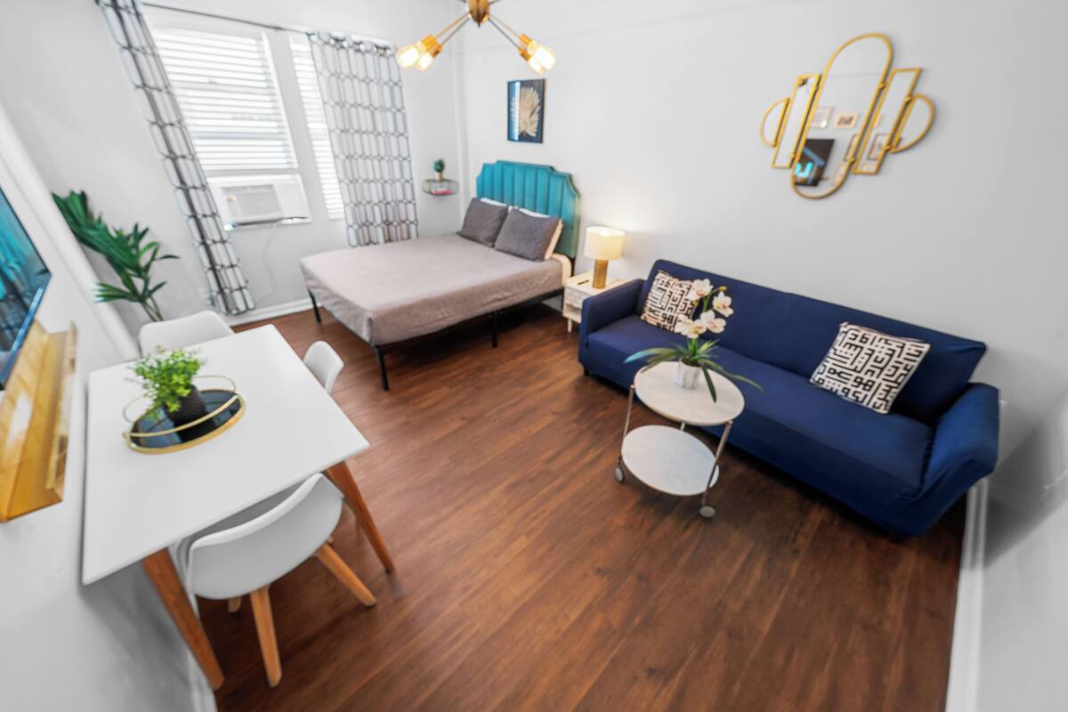 A modern living room with a bed, a blue sofa, a dining table with chairs, and a small round table with a plant.