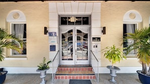 L’entrée d’un hôtel, avec une porte à deux battants, un petit escalier et deux plantes en pot de chaque côté.