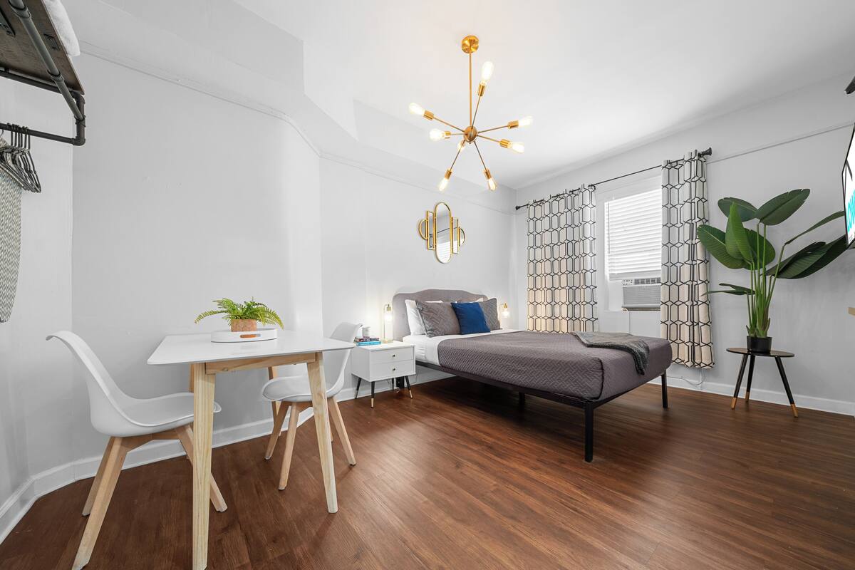 A modern bedroom with a bed, a desk, and a chair. There is a plant on the desk and a potted plant beside the bed.