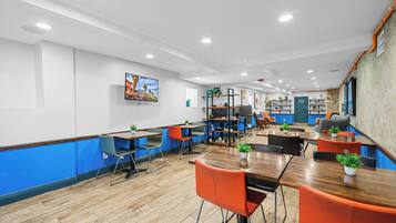 Un comedor moderno con mesas de madera y sillas color naranja, un televisor montado en la pared y estanterías con plantas y objetos decorativos.