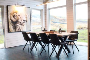 Un espace repas avec une grande table et des chaises, une œuvre d’art encadrée représentant la tête d’un bélier, et une vue sur un paysage montagneux par de grandes fenêtres.