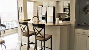 A modern kitchen with two bar stools, a refrigerator, a microwave, and a window overlooking a cityscape. - Beautiful Penthouse Style on Clearwater Beach (Clearwater)