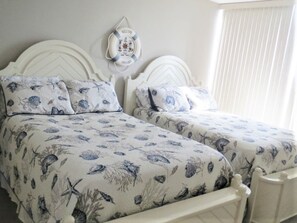 Cama doble con cubierta y almohadas a juego con motivos náuticos, un adorno de flotador en la pared, y una ventana con persianas.