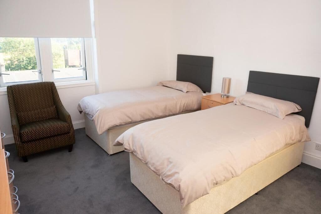 Un dormitorio con dos camas individuales, una silla, una mesita de noche y un ventanal.